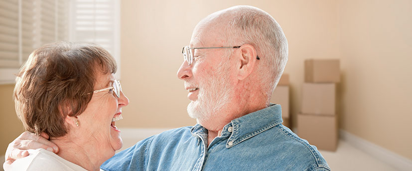 A senior couple, both with glasses are embracing each other and visibly happy in their new space, which is still being moved into and has a few moving boxes in the corner.