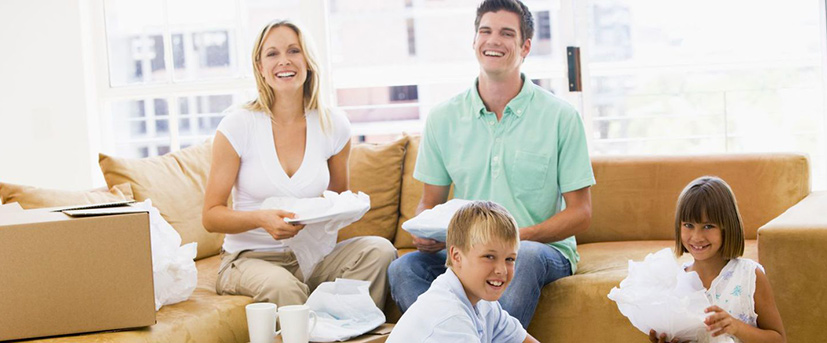 : A family of four is seen unpacking dishes after a move. The mother and father are smiling on a tan colored sectional sofa each holding dishes wrapped in white packing paper and their son and daughter are sitting on the floor also unwrapping dishes.