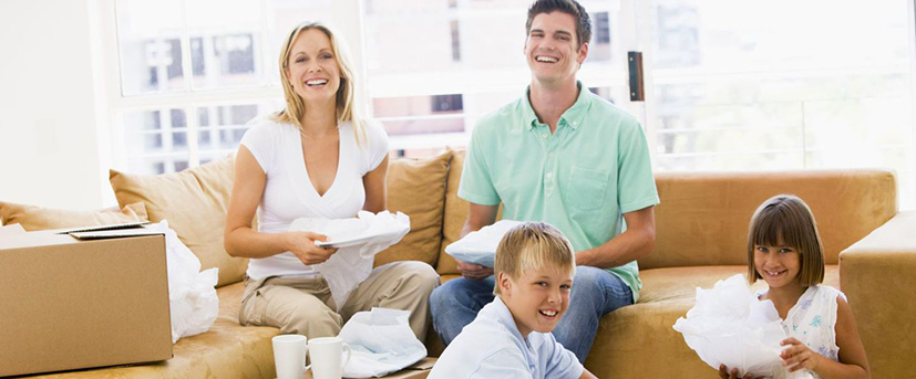 : A family of four is seen unpacking dishes after a move. The mother and father are smiling on a tan colored sectional sofa each holding dishes wrapped in white packing paper and their son and daughter are sitting on the floor also unwrapping dishes. 