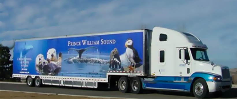 A white semi-truck is seen on the side of the road. The trailer the truck is pulling displays the text “Prince William Sound” on the side with images of otters, a puffin and a whale.