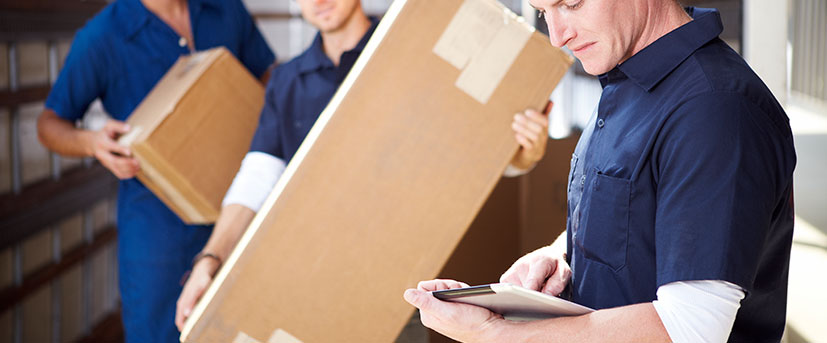 Two out-of-focus movers in full navy-blue uniforms are holding one moving box and standing near a third mover who is checking off a manifest on a tablet.